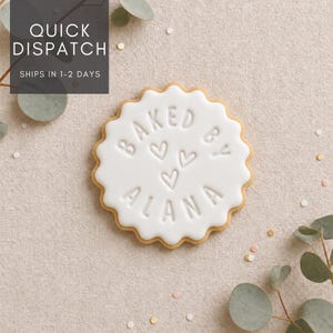 May include: A round, scalloped-edge biscuit with a smooth white icing surface. The text BAKED BY ALANA is embossed in the centre, accompanied by three small heart outlines positioned between the two lines of text.