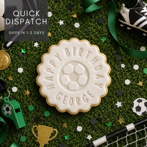 May include: A round, scalloped-edge sugar cookie with white icing featuring a central soccer ball design and the embossed text HAPPY BIRTHDAY GEORGE, surrounded by soccer-themed party decorations on a green artificial grass surface.