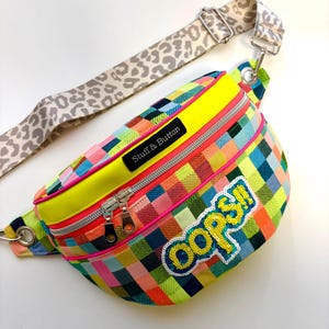 Könnte beinhalten: Bunte Bauchtasche mit einem karierten Muster in Rosa, Blau, Grün und Orange. Die Tasche hat eine neongelbe Oberseite und einen Leopardenmuster-Gurt. Die Texte "Stuff & Button" und "oops!!" sind sichtbar.