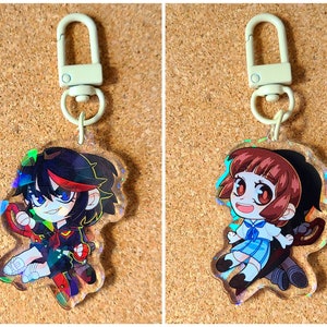 Puede incluir: Dos llaveros iridiscentes con personajes de anime. El llavero de la izquierda presenta a una chica con cabello rojo y negro, vestida con un atuendo negro y rojo. El llavero de la derecha presenta a una chica con cabello castaño, vestida con un atuendo azul y blanco.