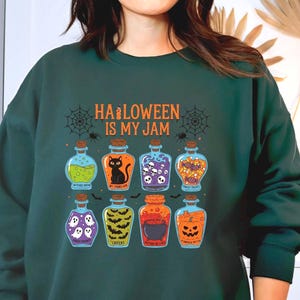May include: Dark green sweatshirt with the phrase "HALLOWEEN IS MY JAM" and illustrations of potion bottles. The bottles contain various Halloween-themed elements like a black cat, ghosts, and a pumpkin.