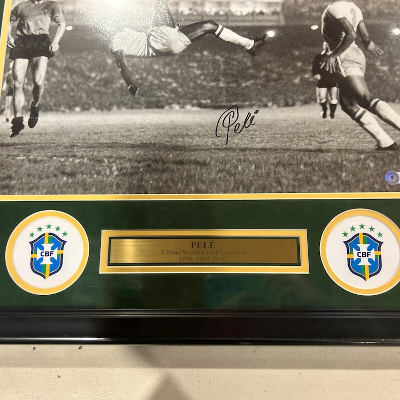 May include: A black and white photo of soccer player Pel&eacute; in mid-air, kicking the ball. The photo is framed with a gold border and a green mat. The mat features two circular emblems with the Brazilian flag and the letters "CBF" in the center. A gold plaque below the photo reads "PEL&Eacute; 3 Time World Cup Champion 1958, 1962, 1970".