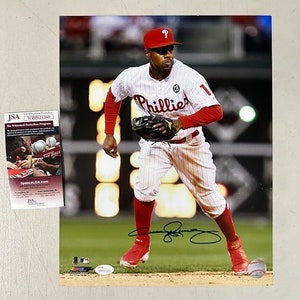 Può includere: Un giocatore di baseball che indossa una divisa bianca e rossa con la scritta "Phillies" sul davanti, tiene un guanto da baseball. Il giocatore indossa un berretto da baseball rosso con una "P" bianca. Il giocatore è in piedi su un campo da baseball.