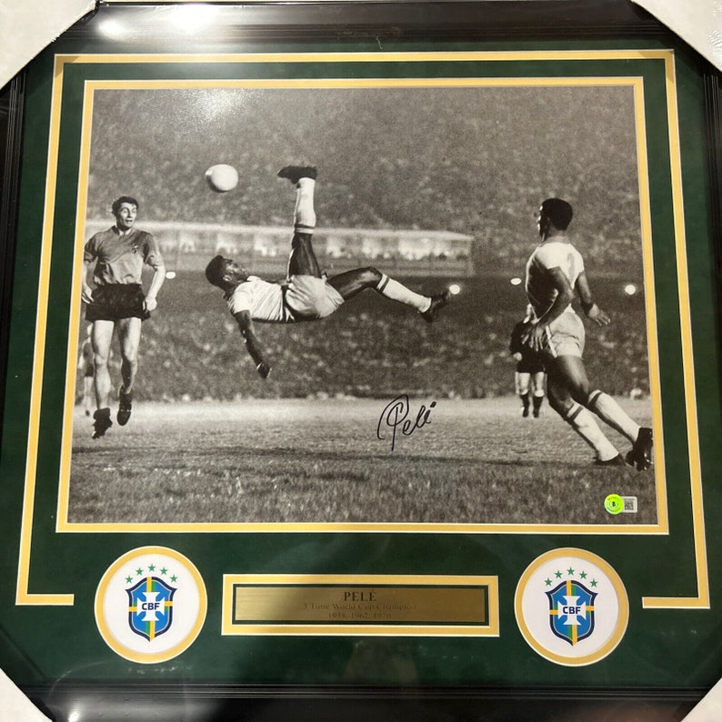 May include: A black and white framed photo of soccer player Pel&eacute; in mid-air, kicking a soccer ball. The photo is signed "Pel&eacute;" in black ink. The frame has a gold border and green matting. The matting features two circular patches with the Brazilian flag and the letters "CBF" in the center.