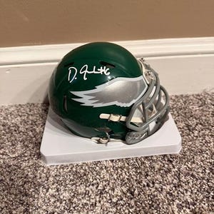 Puede incluir: Un mini casco de fútbol americano verde y plateado con una máscara facial. El casco presenta el logotipo de los Philadelphia Eagles y una firma en tinta blanca. El casco se exhibe sobre un soporte blanco.