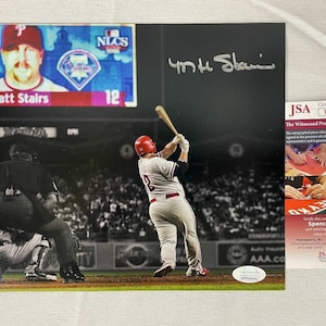 Puede incluir: Una foto en blanco y negro de un jugador de béisbol conectando un jonrón. El jugador lleva un uniforme rojo y blanco y un casco rojo. La foto está firmada por el jugador, "Matt Stairs". La foto está autenticada por JSA, con el número de certificación WPP696944.