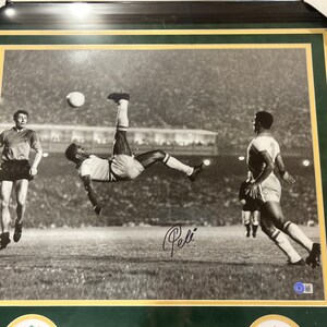 May include: Black and white photograph of a soccer player kicking a ball in mid-air. The player is wearing a white jersey and shorts. The signature "Pel&eacute;" is written in black ink in the bottom right corner of the image.