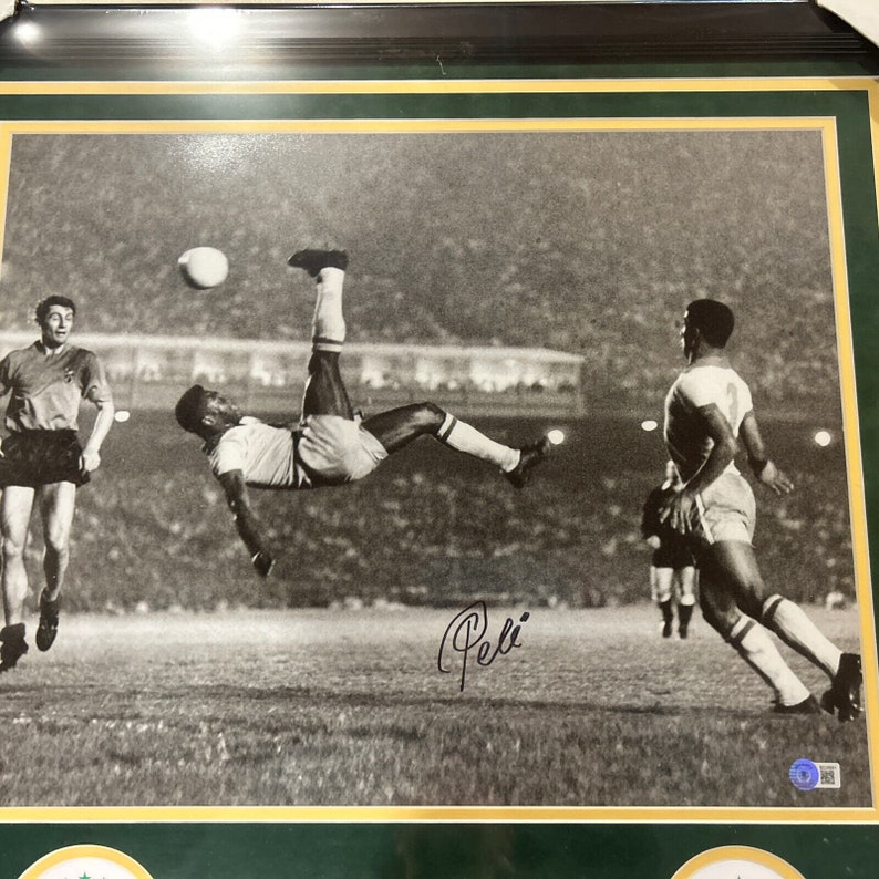 May include: Black and white photograph of a soccer player kicking a ball in mid-air. The player is wearing a white jersey and shorts. The signature "Pel&eacute;" is written in black ink in the bottom right corner of the image.