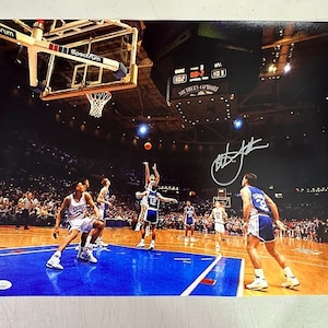 Peut inclure: Une photographie de basketball signée montrant un match de basketball en cours. Les joueurs portent des uniformes bleus et blancs. Le tableau d'affichage indique "00:02" et "103". Le texte "Southern Comfort" est visible sur le tableau d'affichage. La photographie est signée par un joueur à l'encre noire.