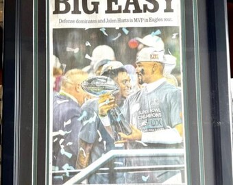 May include: A framed newspaper clipping from The Philadelphia Inquirer with the headline "BIG EASY" and a photo of the Philadelphia Eagles celebrating their Super Bowl LIX victory. The text below the headline reads "Defense dominates and Jalen Hurts is MVP in Eagles rout."