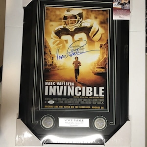 Op de afbeelding: Een ingelijste filmposter voor de film "Invincible" met Mark Wahlberg. De poster heeft een zwarte achtergrond met de titel in witte tekst. De poster is gesigneerd door Vince Papale, de echte persoon waarop de film is gebaseerd. De poster is omgeven door een zwarte lijst met een gouden rand. De poster wordt ook vergezeld door twee kleine, ronde, zilverkleurige tokens.