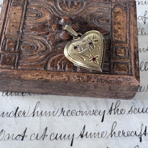 May include: A gold-toned heart-shaped locket featuring an engraved floral pattern with three small red stones set in the center of each flower. The locket has a metal bail at the top and is resting on a dark, carved wooden box with ornate details.