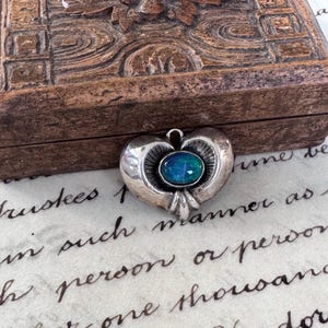 May include: A silver pendant featuring a central oval-shaped iridescent blue and green stone. The metal setting has a smooth, rounded heart-like shape with radiating etched lines surrounding the stone and a small loop at the top for attachment.