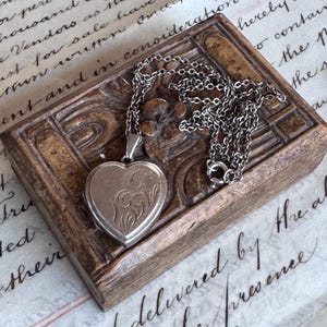 May include: A heart-shaped silver locket pendant on a matching silver chain. The front of the locket features an engraved floral pattern with swirling lines. The metal has a polished finish, and the chain consists of small, interconnected oval links.
