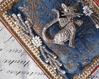 Könnte beinhalten: Silberne Katzenbrosche mit rotem Edelstein auf blauem Stoffhintergrund. Die Katze hat eine Fliege und ist von Perlenakzenten und weißen floralen Verzierungen umgeben. Der Stoff ist mit gold-braunem Zierrat umrahmt.