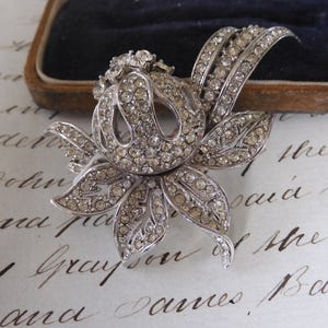 May include: A silver-toned floral brooch, encrusted with clear rhinestones. The brooch features a detailed flower design with multiple petals and a central cluster of stones. The piece is set against a vintage-style background.