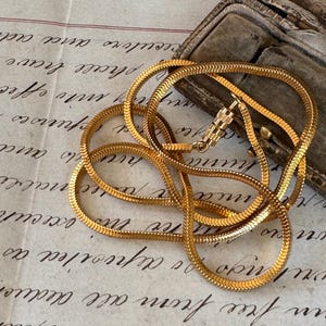 Puede incluir: Un collar de cadena de serpiente de color dorado con un diseño texturizado en forma de escamas. El collar está sobre un papel vintage con escritura manuscrita, junto a un pequeño joyero antiguo abierto.