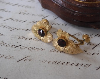 May include: A pair of vintage gold-coloured screw-back earrings. Each earring features a leaf-shaped design with a dark red gemstone centre. The earrings are set against a backdrop of antique script.