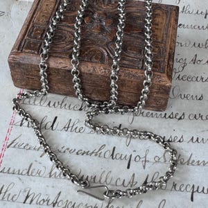 Könnte beinhalten: Eine silberfarbene Metallkette mit runden, ineinandergreifenden Gliedern und einem einfachen Hakenverschluss. Die Kette ist in mehreren Schlaufen drapiert, was die gleichmäßige Textur und die reflektierende Oberfläche der einzelnen Glieder zeigt.