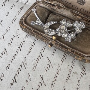 May include: A silver-toned brooch with a floral design, featuring three clusters of sparkling stones and a curved accent. The brooch is displayed inside a vintage, brown leather jewelry box with a velvet interior.