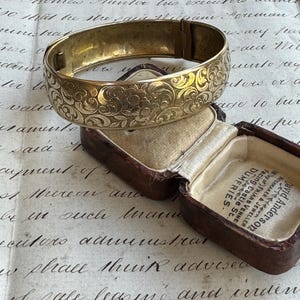 May include: A gold-coloured hinged bangle bracelet with an ornate floral design. The bracelet is resting on a brown leather jewellery box with a cream-coloured interior. The box is open, revealing the interior.