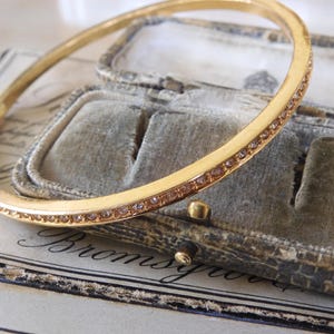 Puede incluir: Un brazalete de oro con una fila de pequeñas piedras transparentes alrededor del borde. El brazalete está sobre una caja de terciopelo marrón con la palabra "Bromsgrove" escrita en ella.