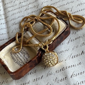 May include: A gold-toned necklace with a round, crystal-studded pendant rests in an open, vintage jewelry box. The necklace has a snake chain design. The box is on a handwritten document.