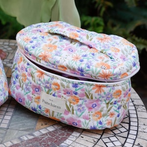May include: A white oval-shaped quilted makeup bag with a floral print featuring pink, orange, and purple flowers. The bag has a zipper closure and a handle on top. The bag has a label that reads "Est. 21 Punchiesss".