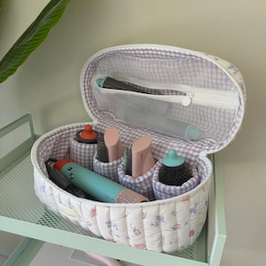 May include: A white quilted oval-shaped travel case with a floral print and a purple and white checkered lining. The case is open and contains a variety of hair styling tools, including a hairdryer, curling iron, and brushes.