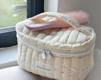 May include: A quilted, cream-colored cosmetic bag with a floral pattern and a matching pink hairbrush. The bag has a white zipper and a small label that reads "Punchiess". The bag is sitting on a light-colored surface.