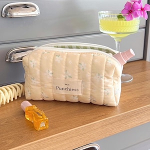 May include: A quilted, pale yellow cosmetic bag with a floral pattern and the text "Punchiess." A small perfume bottle and a cocktail with a pink flower garnish sit nearby. A hair clip is also visible.