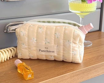 May include: A quilted, pale yellow cosmetic bag with a floral pattern and the text "Punchiess." A small perfume bottle and a cocktail with a pink flower garnish sit nearby. A hair clip is also visible.