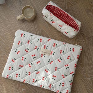 May include: White quilted makeup bag with a red and white checkered lining. The bag has a cherry print and the text "Punchies" on a small tag.