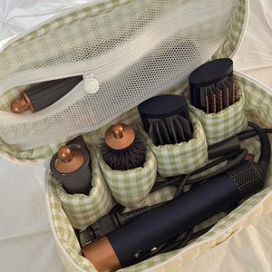 May include: A soft, yellow and green gingham patterned cosmetic bag filled with hair styling tools. The bag contains a hair dryer, brushes, and other attachments. The bag is open, revealing the contents, and is sitting on a white surface.
