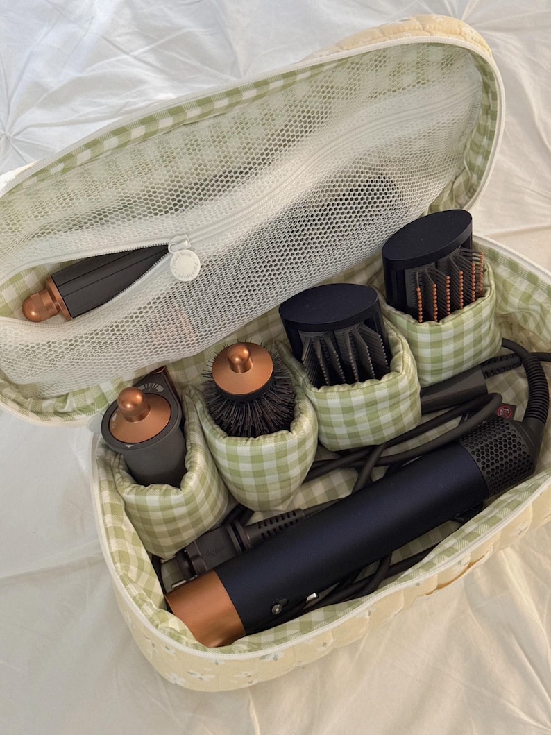 May include: A soft, yellow and green gingham patterned cosmetic bag filled with hair styling tools. The bag contains a hair dryer, brushes, and other attachments. The bag is open, revealing the contents, and is sitting on a white surface.