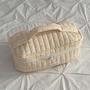May include: A quilted, pale yellow cosmetic bag with a floral pattern and a white zipper. The bag has a handle and the word "Punchiess" is printed on the front. The bag is sitting on a white, textured surface.