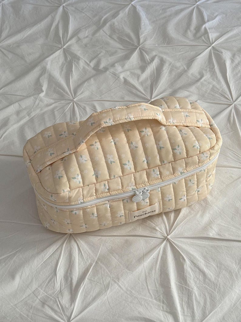 May include: A quilted, pale yellow cosmetic bag with a floral pattern and a white zipper. The bag has a handle and the word "Punchiess" is printed on the front. The bag is sitting on a white, textured surface.