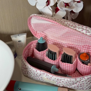 May include: A floral-patterned cosmetic bag with pink gingham pockets holds various hair styling tools, including brushes and a curling wand. A teal and red hair dryer is also visible, along with a tube of Origins face wash.