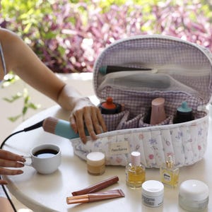 May include: A white and purple quilted makeup bag with a floral print and the text "Punchiesss". The bag is open and contains various makeup and beauty products, including a hairbrush, a bottle of serum, and a tube of lipstick.