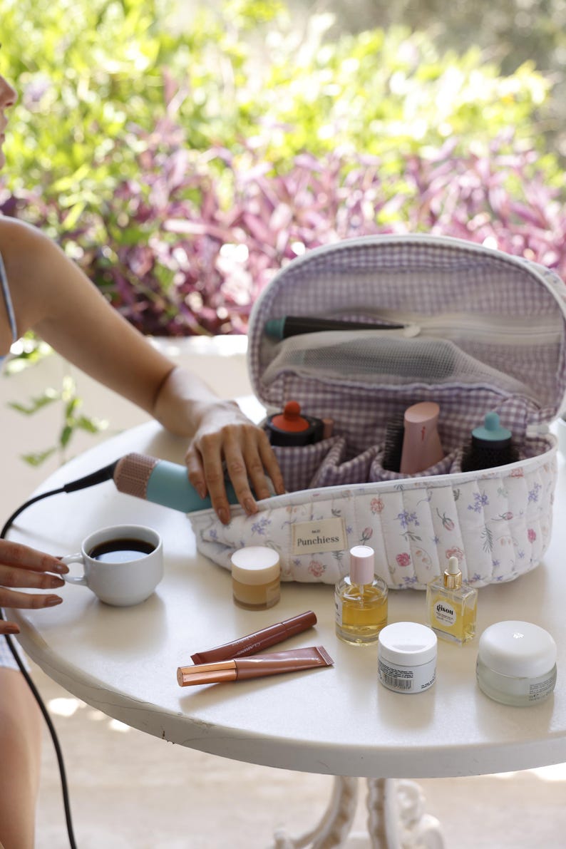May include: A white and purple quilted makeup bag with a floral print and the text "Punchiesss". The bag is open and contains various makeup and beauty products, including a hairbrush, a bottle of serum, and a tube of lipstick.