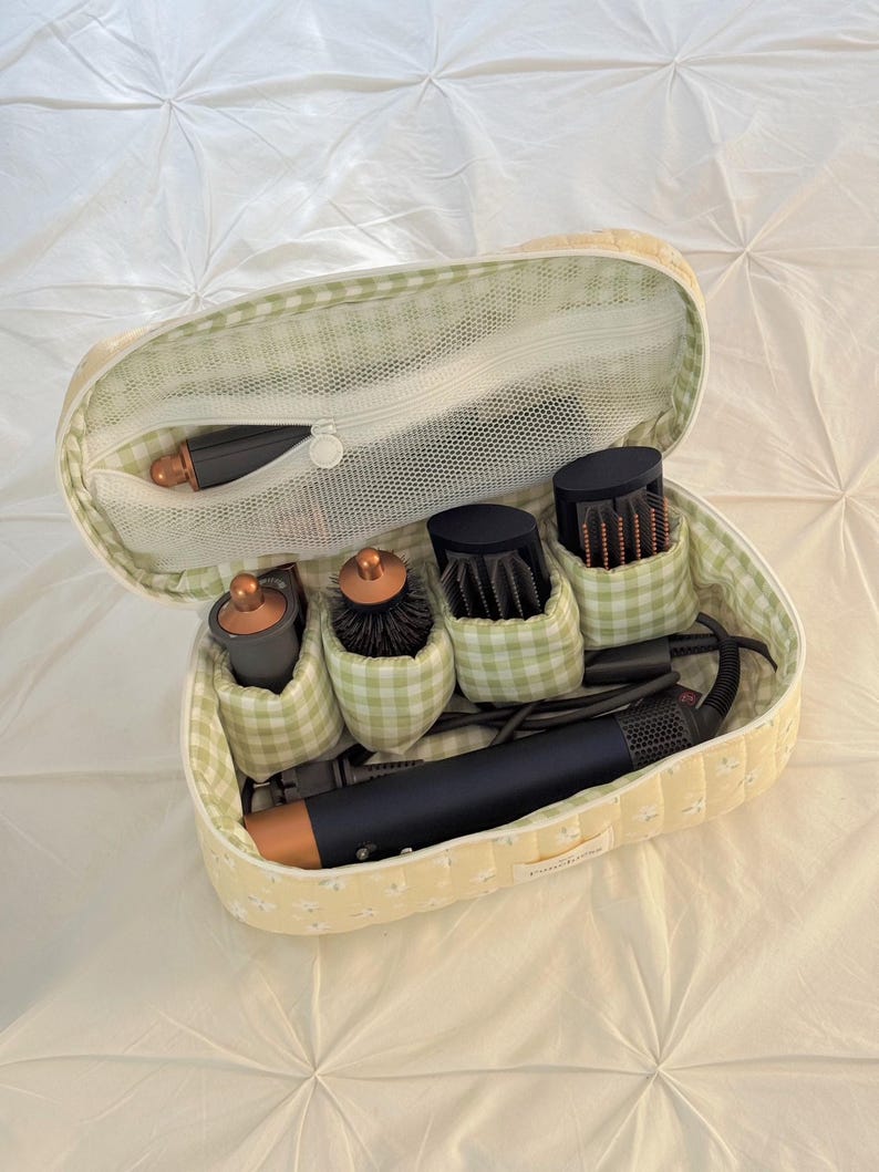 May include: A yellow floral patterned travel case with a green gingham interior, holding a hair styling tool and various attachments. The case is open, revealing a mesh pocket and several compartments for the accessories.