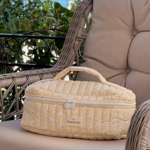 May include: A pale yellow quilted cosmetic bag with a floral pattern and a top handle. The bag has a zipper closure and the word "Punchiess" is printed on the front. The bag is sitting on a beige cushion.