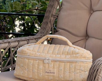 May include: A pale yellow quilted cosmetic bag with a floral pattern and a top handle. The bag has a zipper closure and the word "Punchiess" is printed on the front. The bag is sitting on a beige cushion.