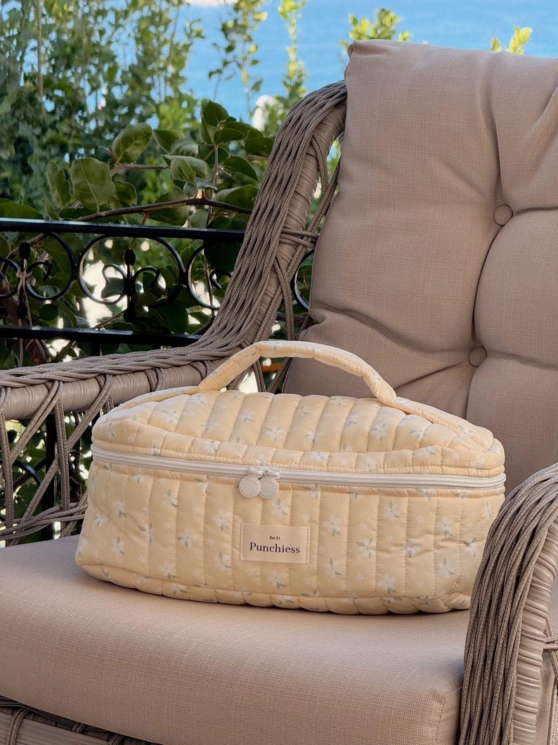 May include: A pale yellow quilted cosmetic bag with a floral pattern and a top handle. The bag has a zipper closure and the word "Punchiess" is printed on the front. The bag is sitting on a beige cushion.