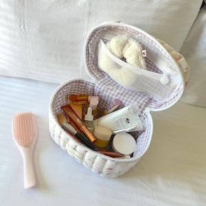 May include: A white quilted heart-shaped makeup bag with a floral print and a pink and white gingham lining. The bag is open and filled with various beauty products, including a hairbrush, a bottle of perfume, and a tube of lipstick.