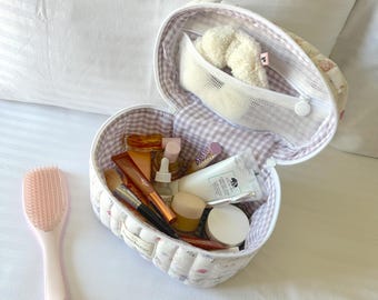 May include: A white quilted heart-shaped makeup bag with a floral print and a pink and white gingham lining. The bag is open and filled with various beauty products, including a hairbrush, a bottle of perfume, and a tube of lipstick.