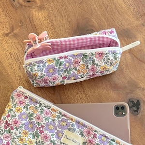 May include: A floral print zippered pouch with a pink and white gingham lining. The pouch is open and has a pink hair clip inside. The pouch has the word "Punchless" printed on the front.