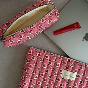 May include: A pink quilted makeup bag with a zipper closure and a floral print. The bag is open and the inside is lined with a cream-colored fabric. The bag is sitting on a gray couch next to a laptop and a red tube of lip balm.