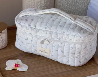 May include: A white quilted cosmetic bag with a blue floral pattern and a top handle. The bag has a zipper closure and a small label that reads "Punchiess". An orchid flower is on the wooden surface.