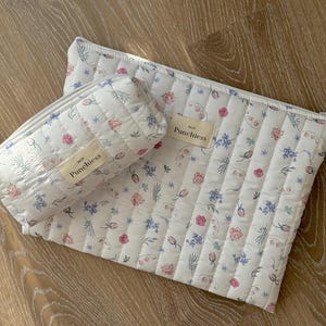 May include: Two white quilted cosmetic bags with a floral pattern of pink and blue flowers and green leaves. One bag is rectangular, the other is cylindrical. Both have a small label that reads "Punchless".